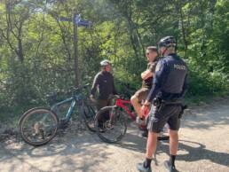 Zwei Radpolizisten, in der Mitte ein Fahrradfahrer, Hintergrund Wald
